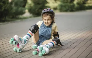 Cute little girl in protective equipment and rollers sitting on walkway