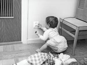Dangerous in the 1960s. A child is sitting on the floor and is curious about the electrical outlet in the wall. She holds a plug in one hand and fingers the outlet with the other