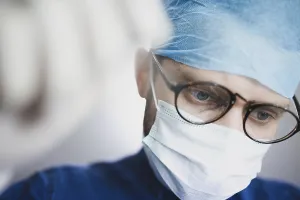 Doctor in a medical setting with a focused expression wearing glasses and a surgical cap