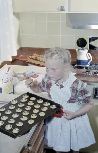In the early 1960s. A girl nicely dressed in an apron puts raisins on top of the almost ready to bake cookies