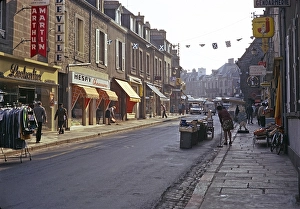 Eine Reihe von Geschaeften mit Menschen, die die zum Marktplatz fuehrende Strasse buersten, vermutlich in Quimper, Bretagne, Frankreich, 1974