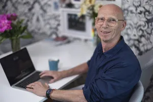 Elderly, bald man with a friendly smile sitting in his living room at a table with flowers and a cup of coffee