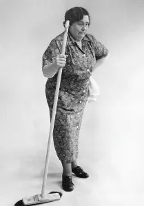 Elderly cleaning lady looking worn and slightly forward as if she has a pain in her back. She does not look very happy standing there leaning against a brush. 1940s