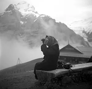 An elderly woman sits in the mountains on a wooden bench and examines the landscape through binoculars