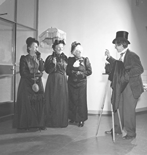 Three elderly women dressed in dresses from the turn of the century pose in front of a photographer with a camera