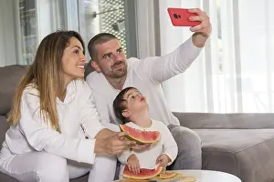 Family with baby boy taking a selfie eating watermelon sitting on the sofa at home