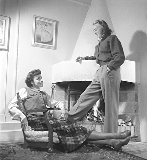 Fashionable in the 1940s. Two women in a room where with a fire burning in the fireplace. One sitting in an extraordinary looking armchair that has no legs. They are both wearing the typical clothes and fashion of the 1940s decade