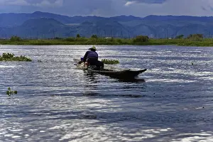 A fisherman uses a paddle on a calm lake surrounded by mountains and clouds
