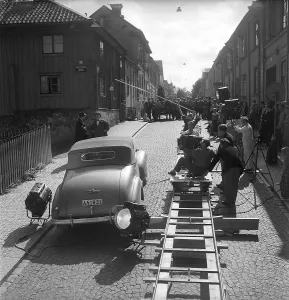 Foer min heta ungdoms skull 1952. Drama film directed by Arne Mattsson. The picture shows filming in Uppsala. The film camera is mounted on a moving platform on rails that follows the actors and the car when it is moving
