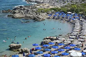 Well frequented beach with many blue parasols and swimmers in the emerald green sea under the midday sun