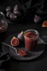 From above fresh fruit jam in small jar and on spoon with full and cut figs on plate at black kitchen table