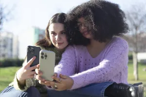 Two friends sitting on grass, taking selfies with their smartphones