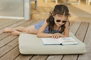 Girl with sunglasses sits on cushion on wooden terrace and reads a book