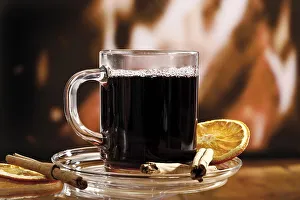 Glass of mulled wine garnished with cinnamon sticks and a dried orange slice in front of a fireplace