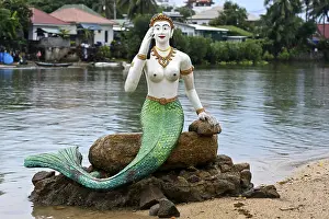 Greeting mermaid statue with white upper body and green tail fin sits on a stone on the shore