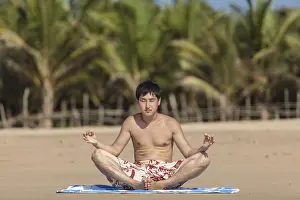 The guy of Asian appearance practices yoga on a beach against palm trees