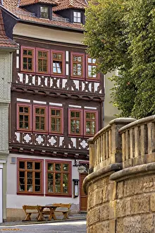 Framed Print : A half-timbered house with red window frames next to a stone wall in a historic town