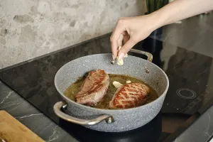 A hand adds garlic to sizzling duck steaks in a pan on the induction stovetop