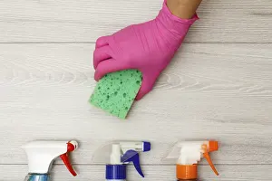 Hand in pink rubber glove holding sponge with bottles of detergent on wooden boards