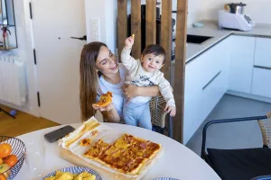 Happy mother holding her baby while eating pizza in a bright