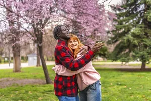Happy multi ethnic couple taking a selfie with smartphone while hugging in a park full of blooming trees during spring
