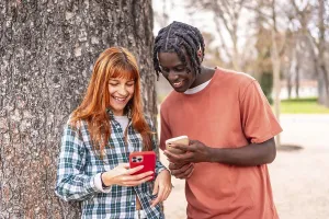 Two happy multi ethnic students sharing funny content on their mobile phones while leaning on a tree in a university