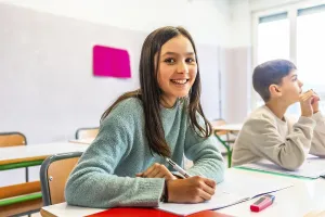 Happy schoolgirl writing on notebook during lesson in classroom with schoolmate thinking in background