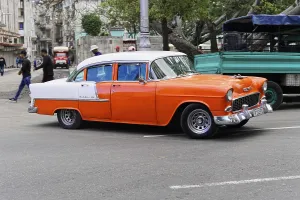 Havana, Cuba, Central America, An orange and white vintage car drives on a road past some trees and buildings