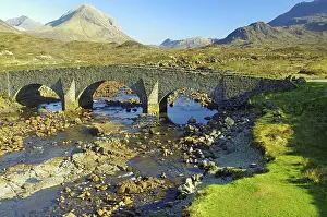 Historic stone bridge over a shallow river with mountains in the background