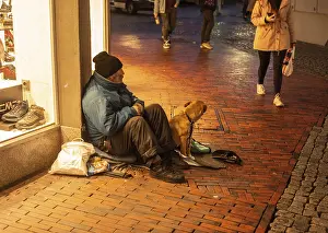 Homeless man with a dog sits in a shopping mall as people walk by in Husum