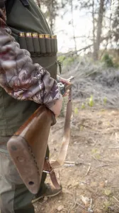 Hunter gripping a shotgun and wearing an ammunition belt while navigating through the dense forest