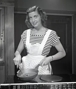 In the kitchen 1940s. A young woman smiles at the camera holding a frying pan and a whisk in her hands