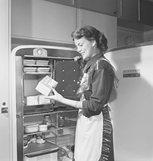 In the kitchen 1950s. Interior of a kitchen and a young woman standing at the Leonard refrigirator with it's door open, showing the cans and bottes of different food standing on the shelfes