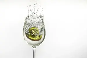 A kiwi falls into a wine glass and creates splashing water against a white background