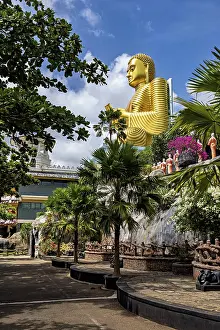 Large golden Buddha statue outdoors surrounded by trees and other sculptures under a blue sky