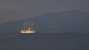 A large sailing ship sails in calm seas against a mountain backdrop at dusk