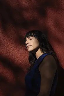 A Latin woman leans against a red wall with her eyes closed