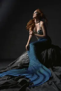Full length of young redhead female wearing bra and blue mermaid tail sitting on rock against black background
