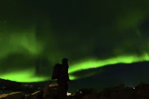 A lone figure silhouetted against vibrant green aurora beneath a star-lit sky