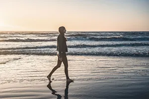 A lonely woman at the beach walking barefooted by the shore at sunrise