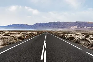 long straight empty asphalt road through arid landscape against ocean and mountain range on Lanzarote