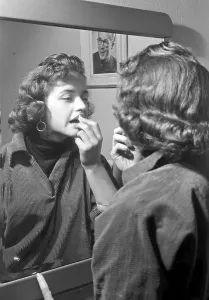 Makeup in the 1950s. A young woman is standing in front of a mirror applying lipstick