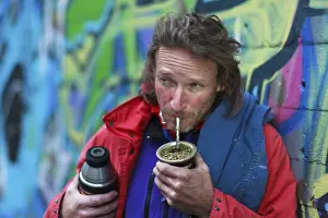 A man with a beard in colourful urban surroundings drinks mate from a cup