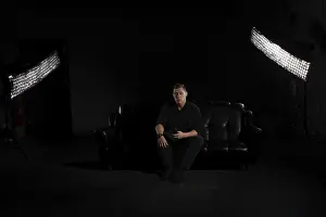 Man in black suit sits on a black couch with a phone