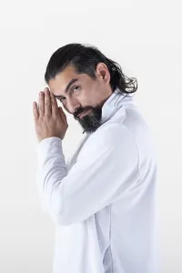 A man dressed in white clasping hands while looking at camera over white background