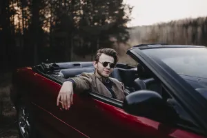 Man smiles while sitting in a red convertible in a forest setting during dusk