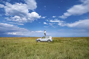 A man stands on a car in the centre of a vast landscape under a blue sky