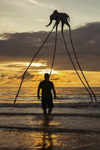 A man stands in the surf facing a surreal elephant sculpture with long legs