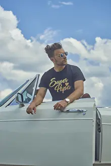 Man in sunglasses leaning on a white convertible car with a cloudy sky backdrop
