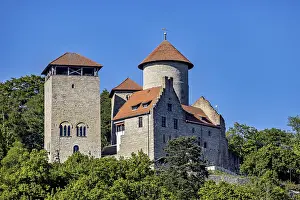 A medieval castle with two towers against a clear blue sky and surrounded by green trees